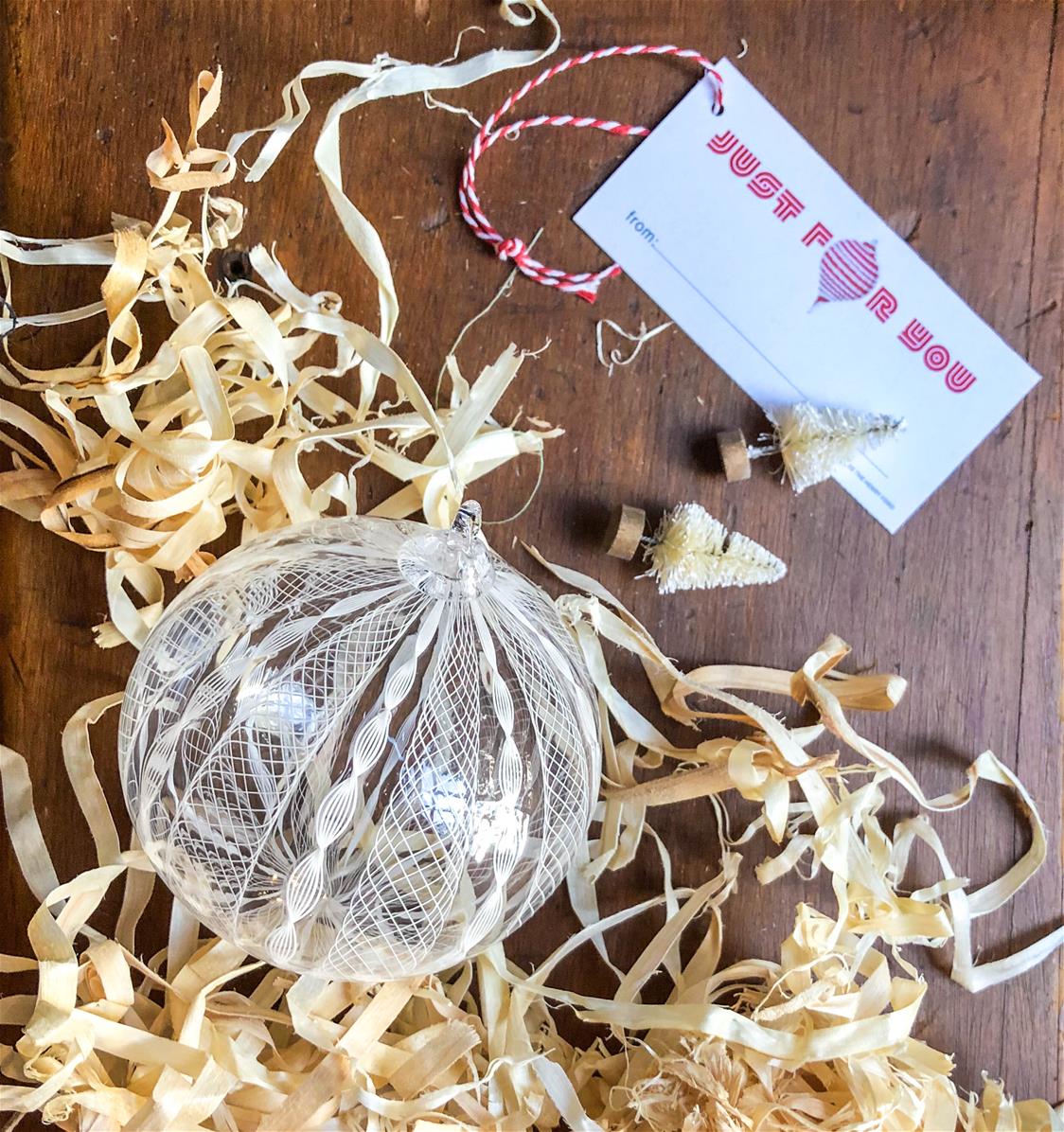 clear and white glass ornament, sitting on table with excelsior, tiny Christmas trees, and a gift tag