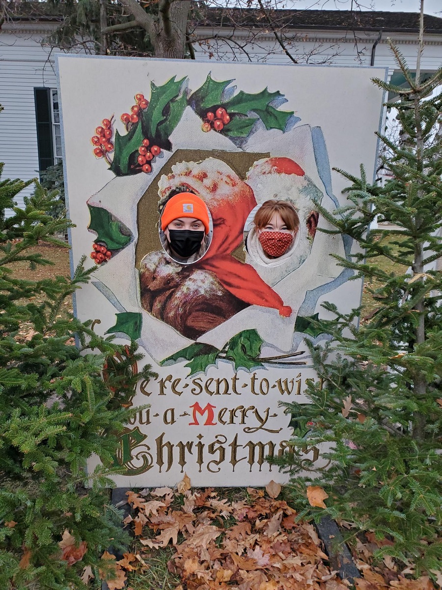 Two people stick their faces through holes in a life-size holiday postcard among evergreens