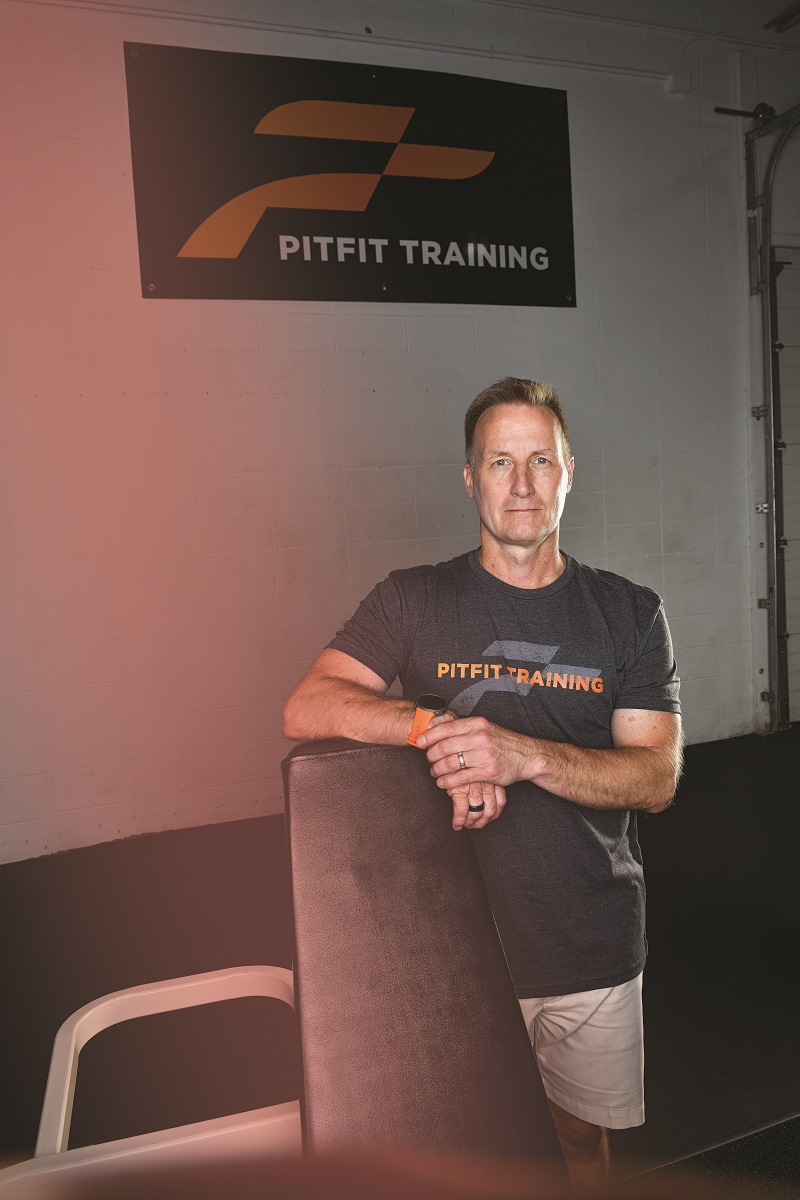 Jim Leo of PitFit Training Man in t-shirt and shorts stands under a sign reading "PitFit Training"