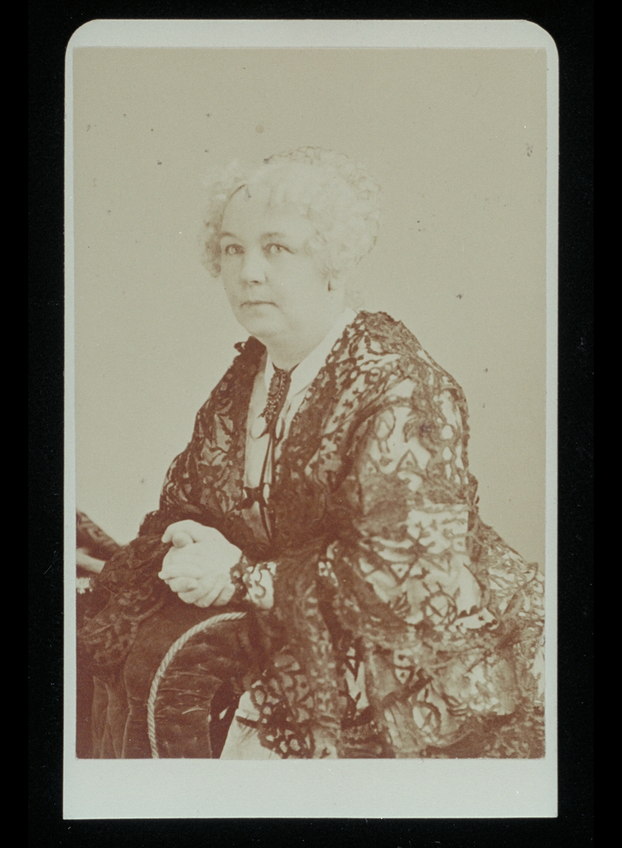 Black-and-white portrait of woman with light-colored hair and clasped hands wearing elaborate lace dress.