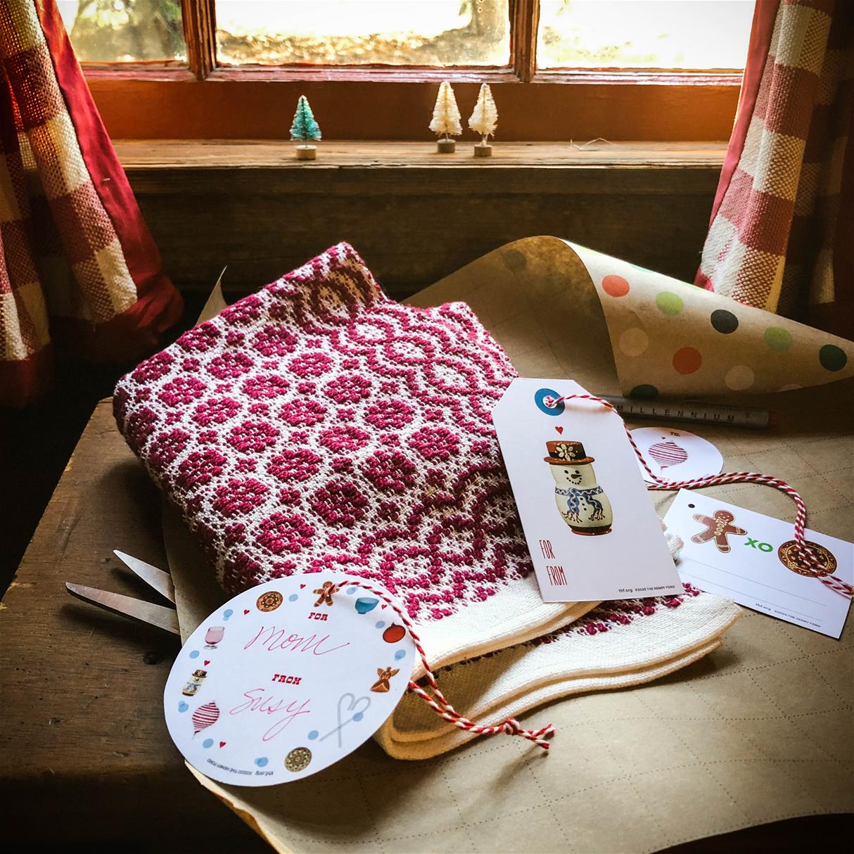 Red-and-white woven placemat on a piece of gift wrap with gift tags; window with red-and-white checked curtains and tiny Christmas trees on windowsill in background