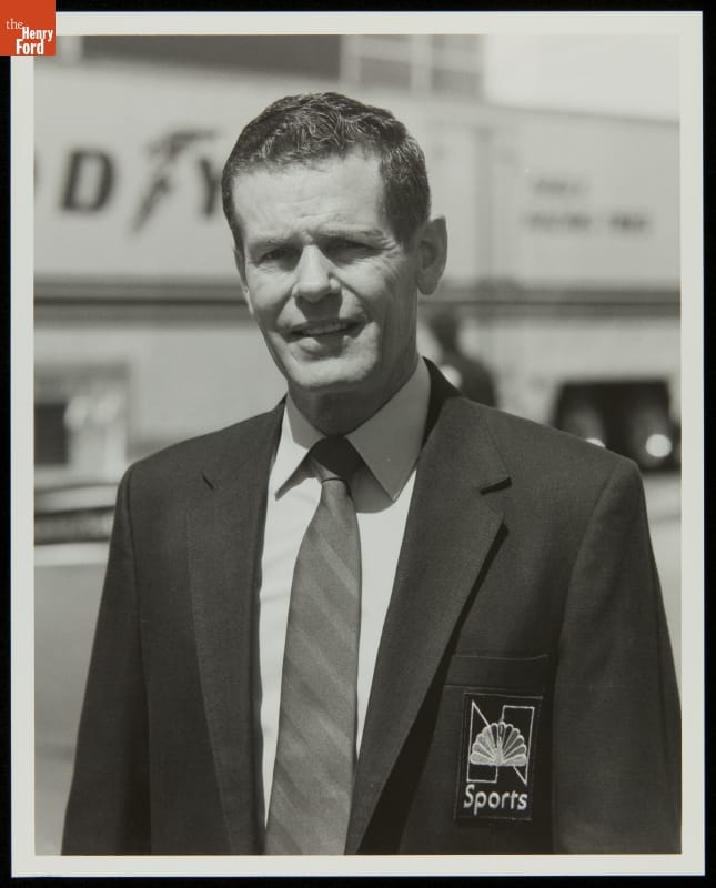 Man in suit and tie with NBC peacock logo on pocket looks at camera