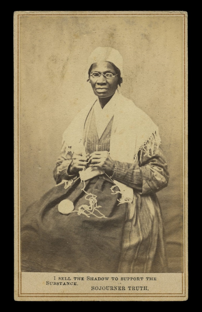 Black-and-white image of dark-skinned woman in shawl and cap knitting and looking at camera; also contains text