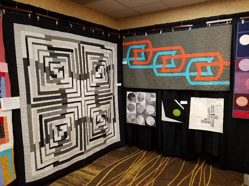 Quilts, mostly shades of black, white, and gray, hanging on a black backboard