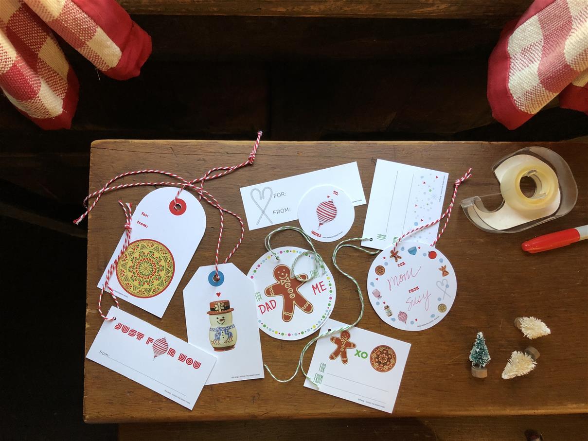 Various shapes and designs of holiday labels on a wood table with tape, scissors, and tiny Christmas trees