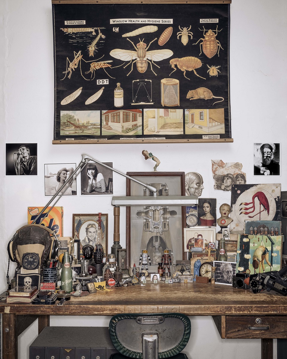Desk littered with models, framed images, knickknacks, and more, with even more items hanging on wall above desk