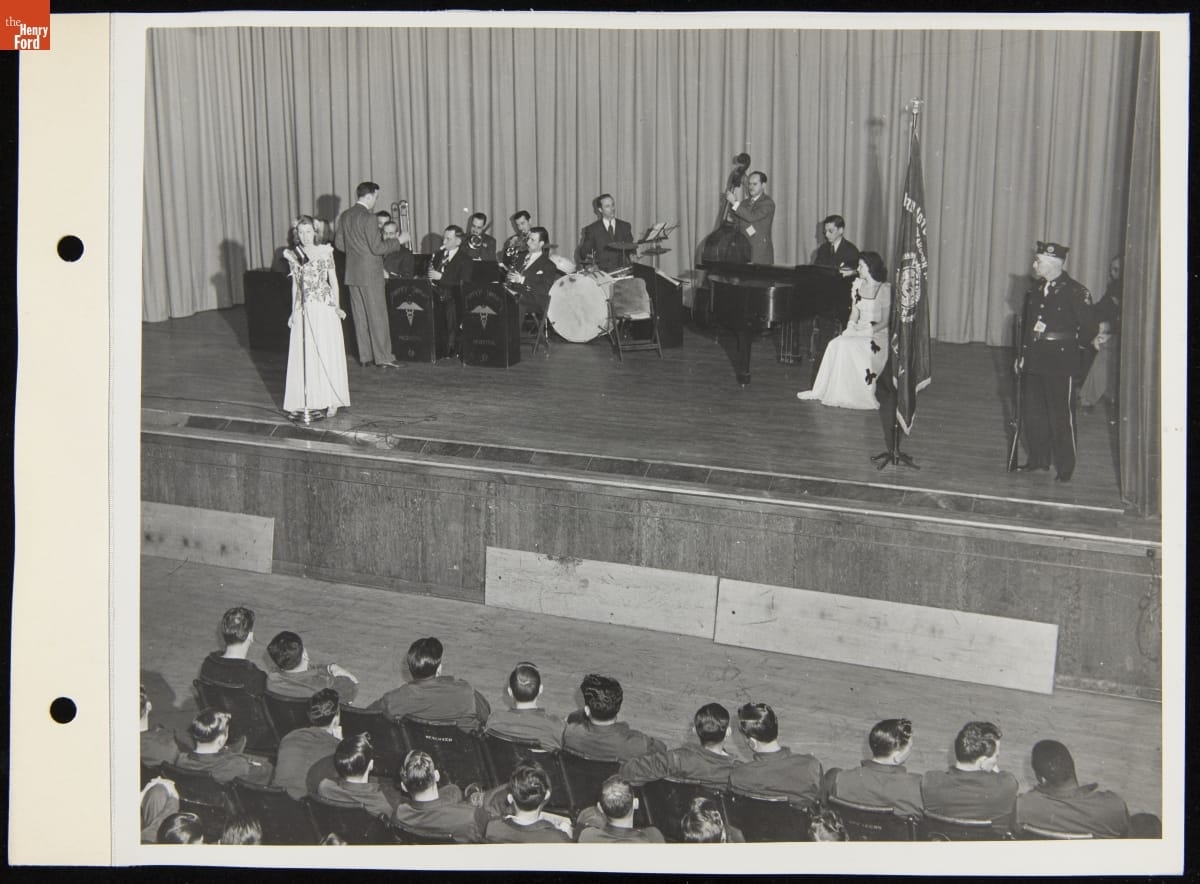 Men with instruments and two women, one at a microphone, on a stage with a soldier in front of an audience