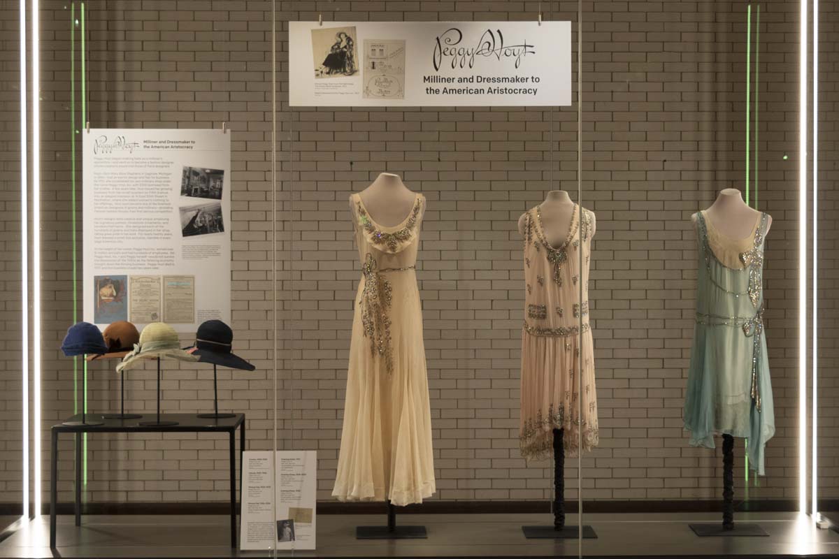 What We Wore exhibit, featuring Peggy Hoyt Large glass display case with three dresses, four hats, and signage with text and images