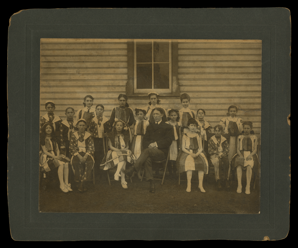 Matted black-and-white photo of group of children wearing stars and stripes sitting and standing with a man in a suit in front of a wooden building