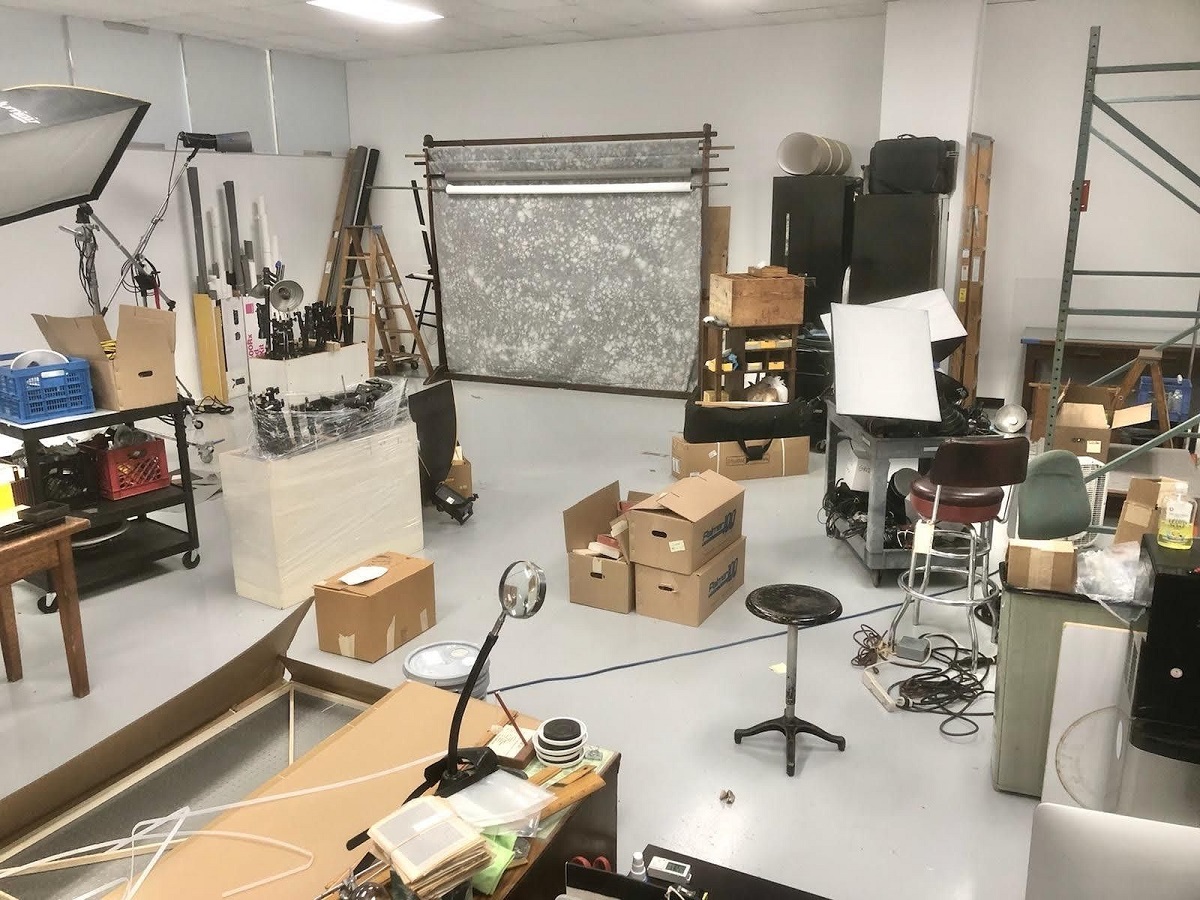 Room cluttered with boxes, furnishings, and other items, with gray and white mottled backdrop in background