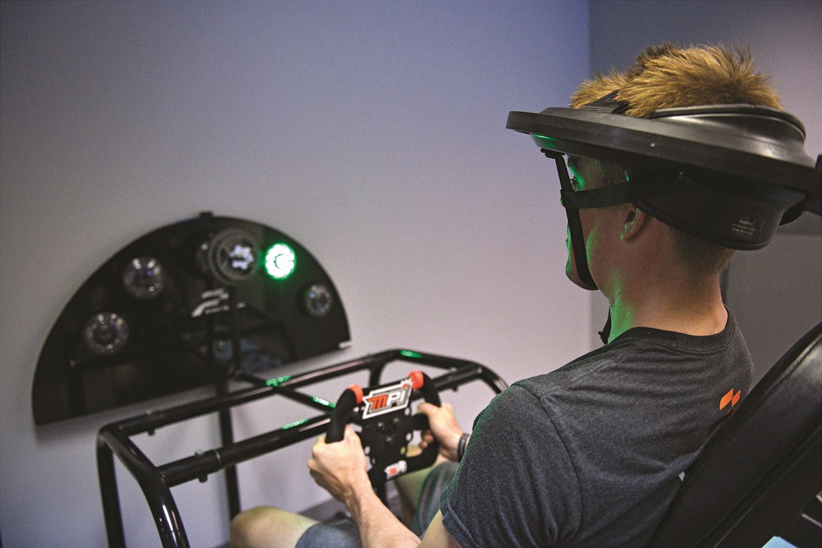 Man wearing large oblong headpiece sits holding what looks like a video game controller in front of a digital dashboard