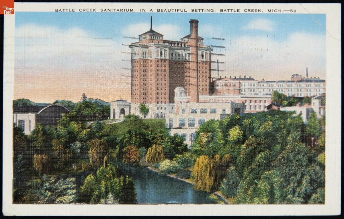 Large white and red-brick building by water, with trees and foliage in front