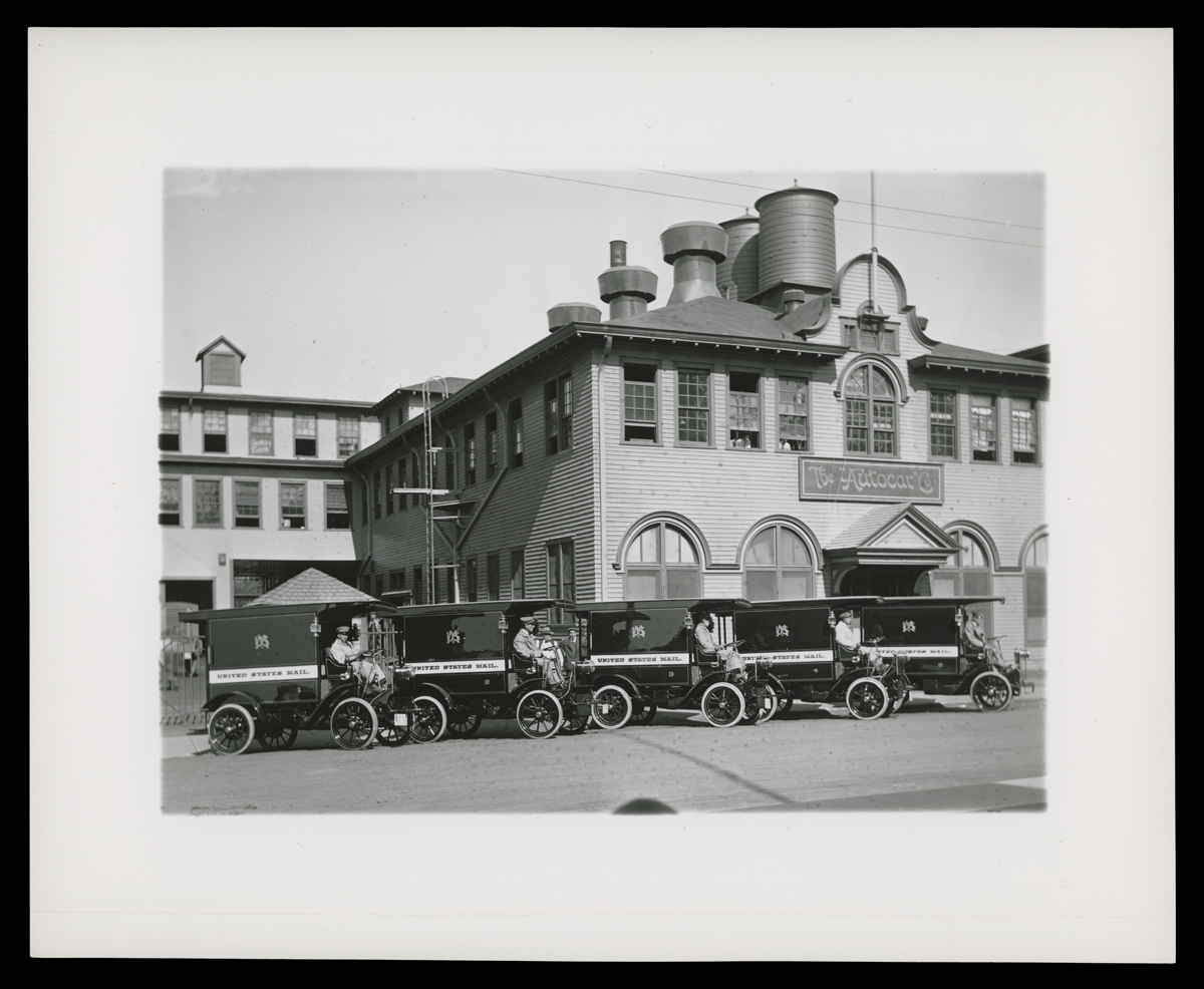 Black-and-white photo of small delivery trucks lined up in front of a large brick building