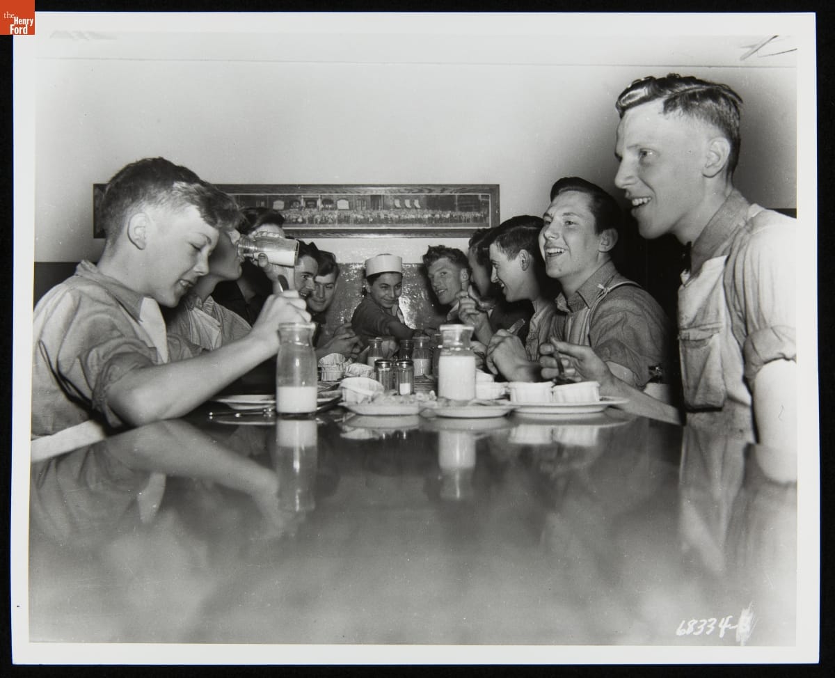Group of boys crowd at one end of a dining table with plates and milk bottles on the table in front of them