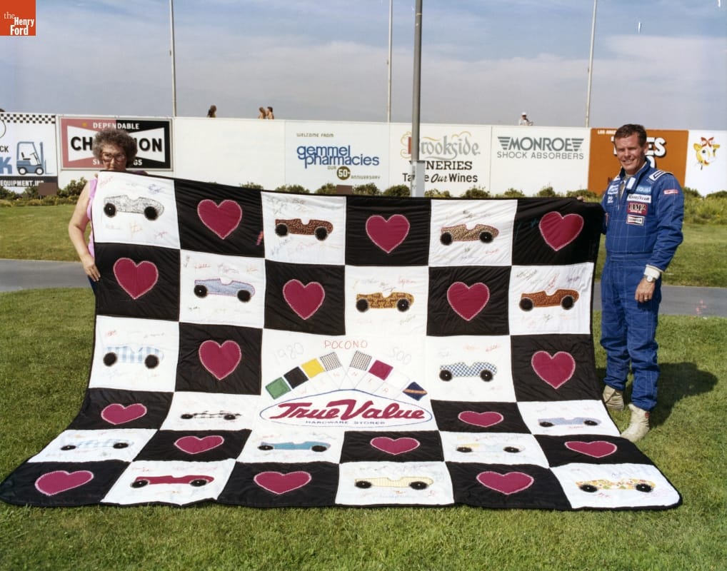 Bobby Unser with Quiltmaker Jeanetta Holder at Pocono in 1980 Man in blue jumpsuit and woman with curly hair wearing tortoiseshell glasses hold corners of a quilt spread out on the grass
