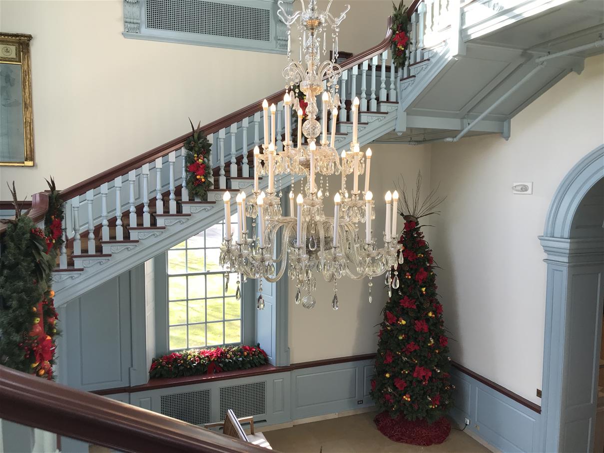 White-and-light-blue room with staircase and elaborate chandelier, decorated with Christmas trees and greenery