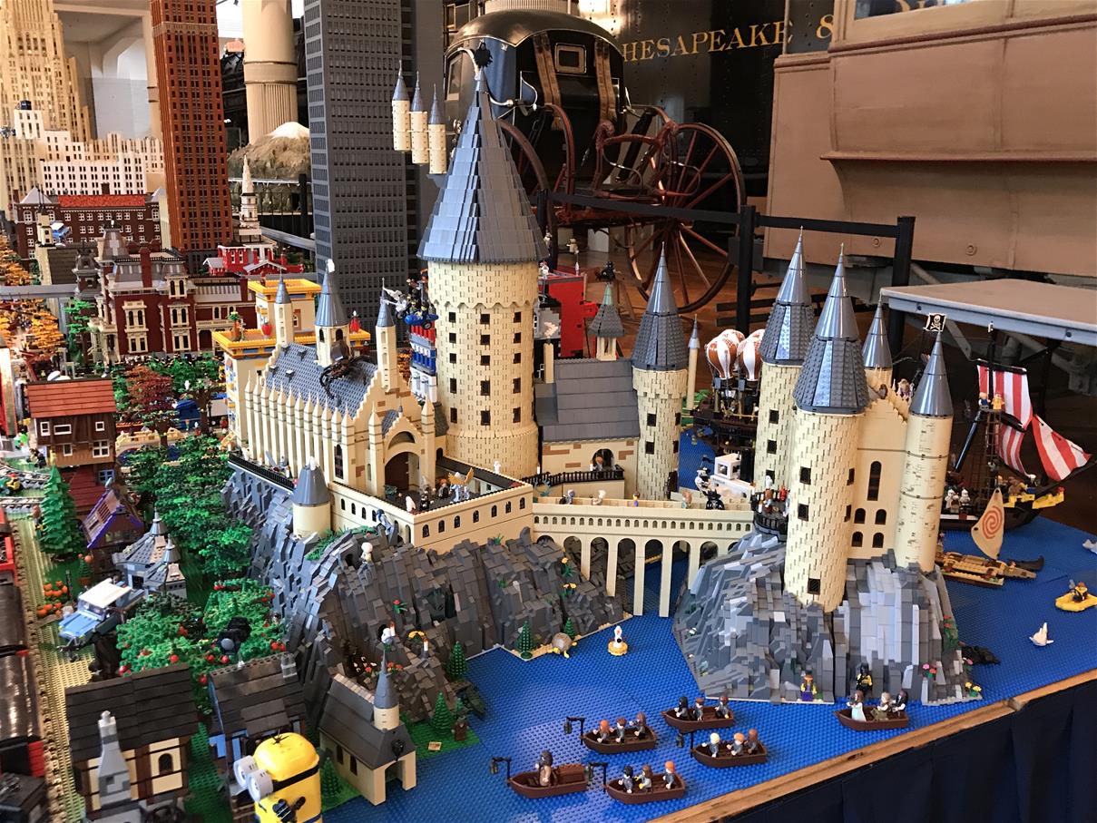 Extensive LEGO layout with boats and castle in foreground, buildings in background, and unrelated museum artifacts behind the layout