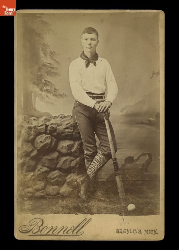 Black-and-white photo of man in early baseball uniform holding baseball bat, leaning on a stone wall in front of a backdrop