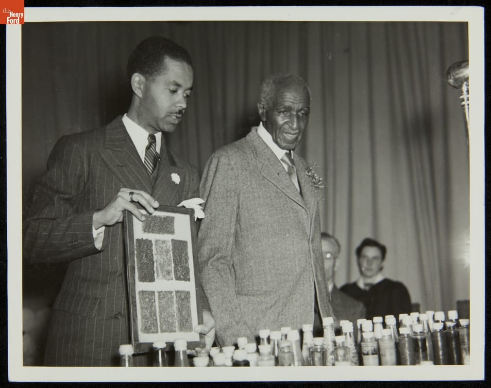 Austin W. Curtis Assisting George Washington Carver during Lecture at Starr Commonwealth for Boys School, Albion, Michigan, 1939 Black-and-white photo of two men in suits standing at table filled with bottles; one man holds a small display board
