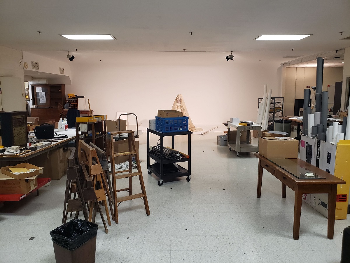Room filled with furniture, stacked ladders, carts, sawhorses, and a wedding dress on a mannequin in the background
