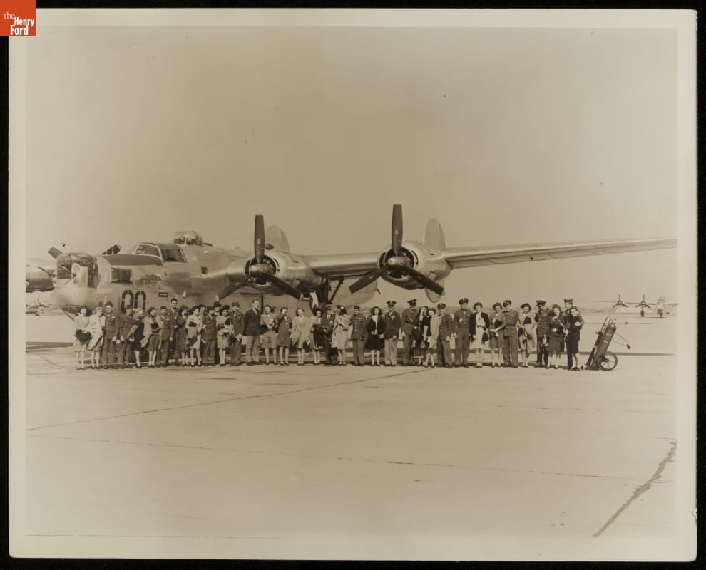 Large group of men and women pose in front of airplane on tarmac