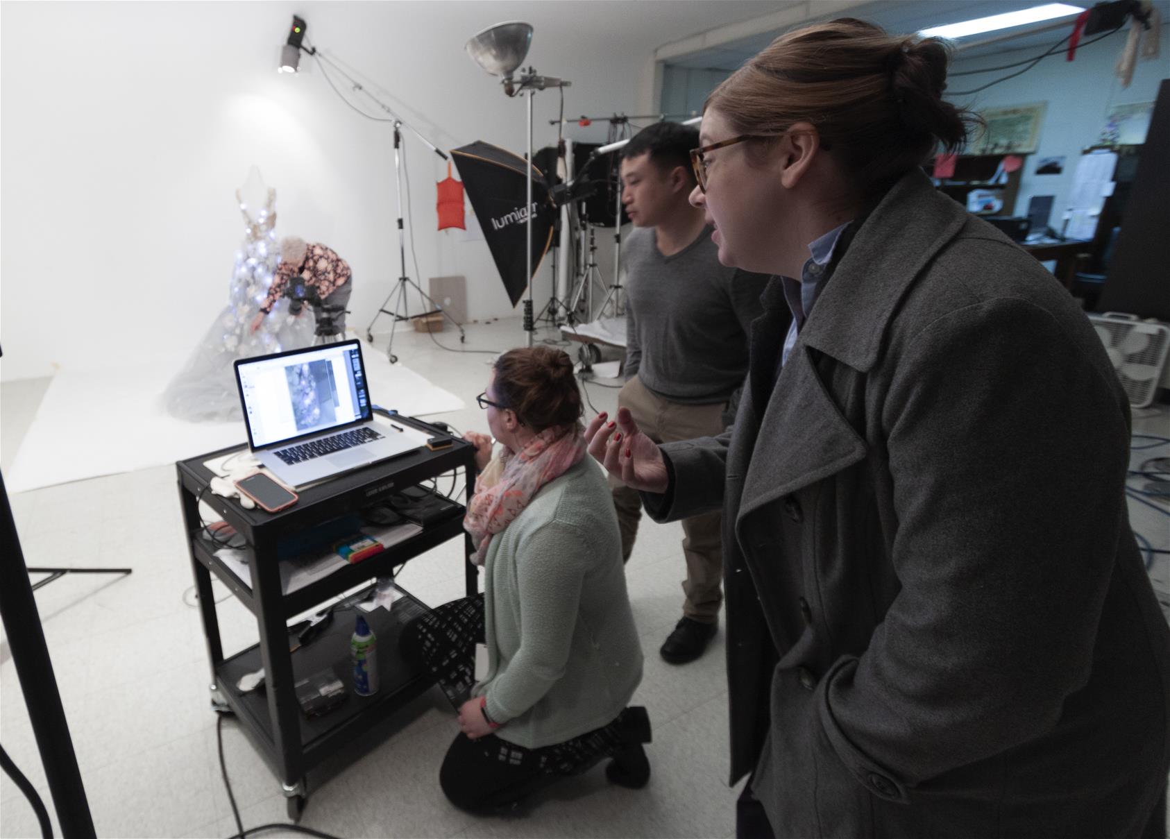 Three people around a computer on a cart, with photography equipment and a woman adjusting a dress on a dress form in the background