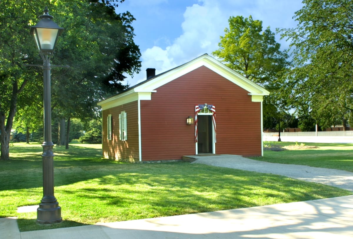 Small red wooden building sitting on a greenspace among trees, sidewalks, and streetlamps