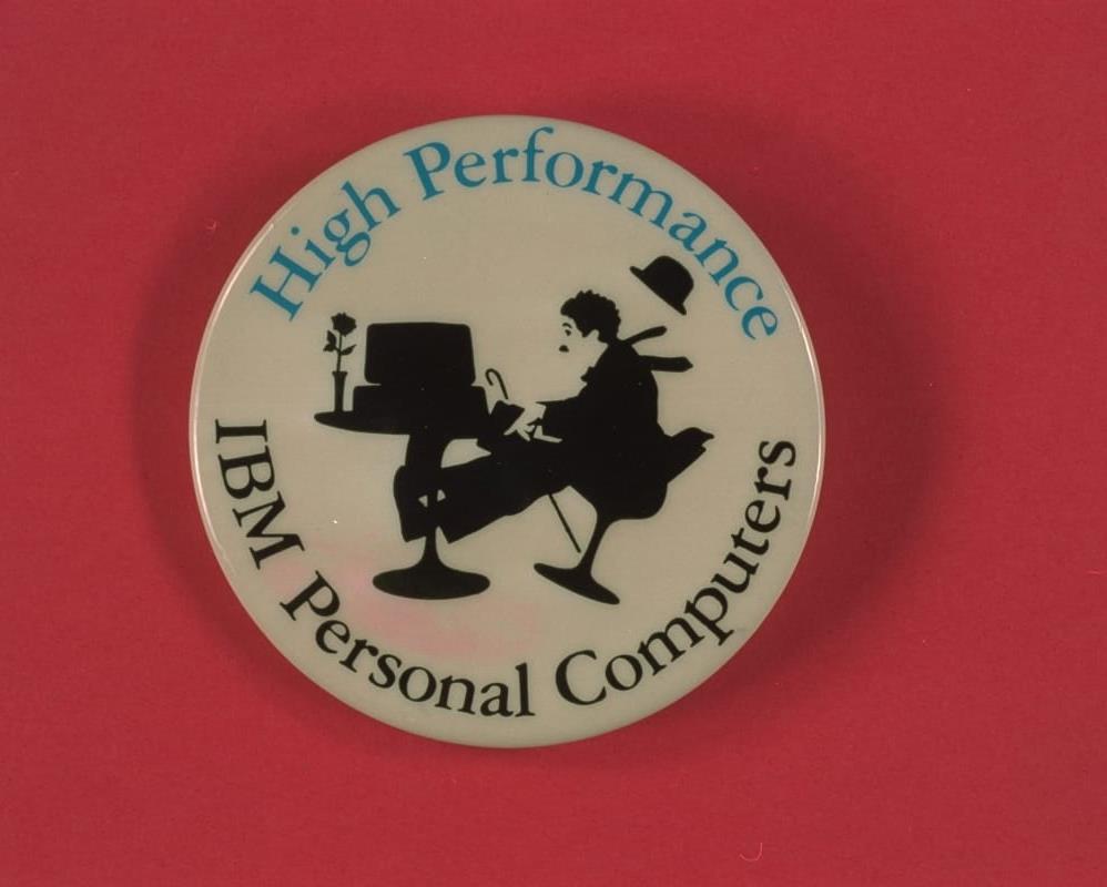 White button with silhouette image of Charlie Chaplin sitting in a chair that appears to be falling backwards at a table that holds a computer and vase of flowers; also contains text