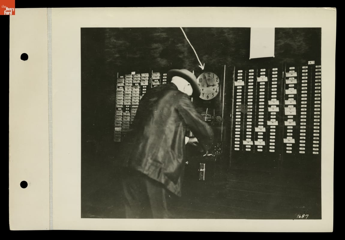 Man in suit and hat slightly leaning over towards a clock and wall with many slots for cards