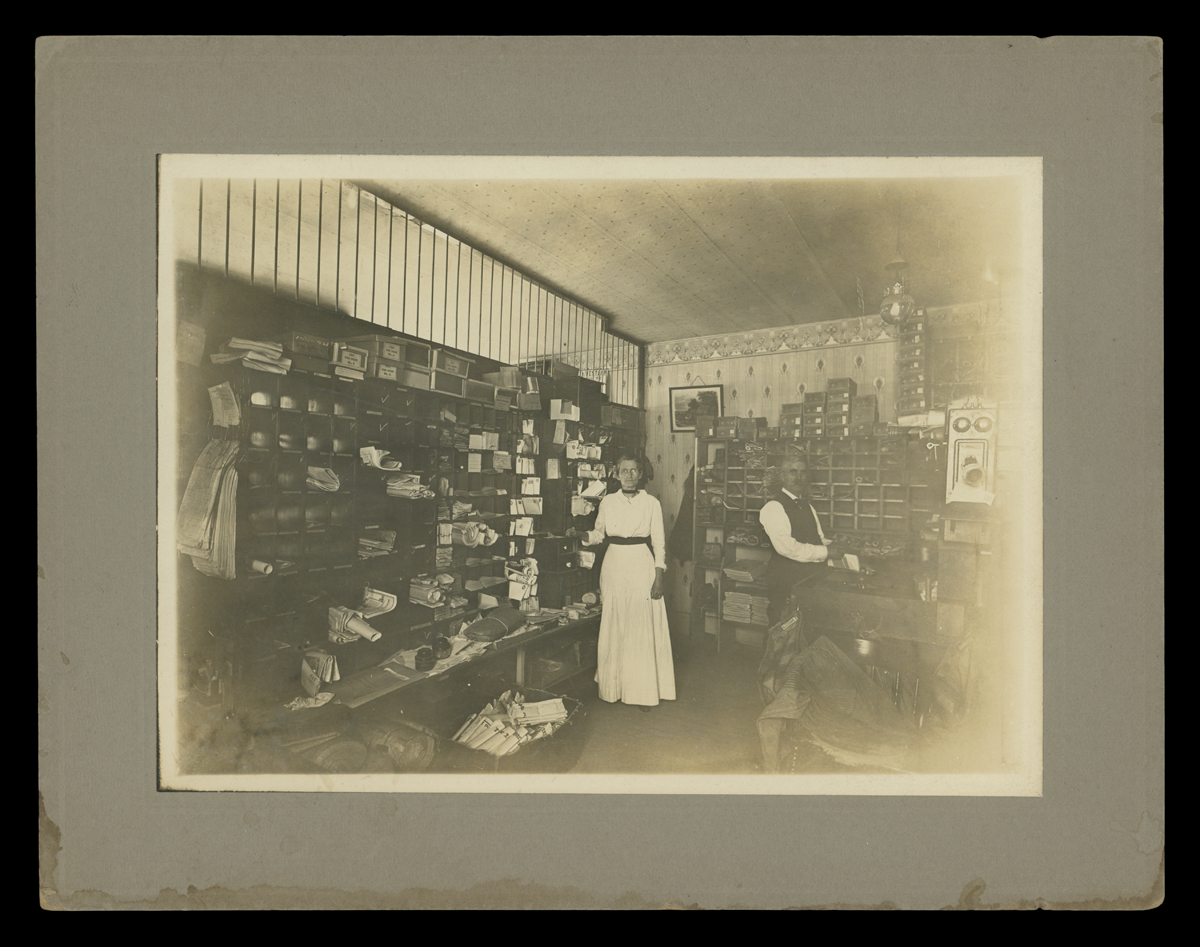 Matted black-and-white photo of a man and woman in a room filled with mail bins and cubbies with items sorted into them