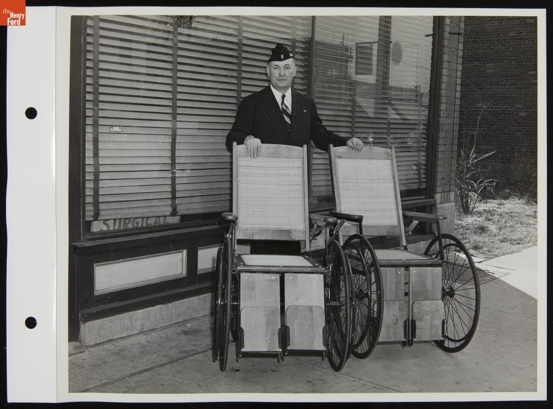Man in suit and military-looking cap stands with hands on two wheelchairs in front of a window with closed blinds