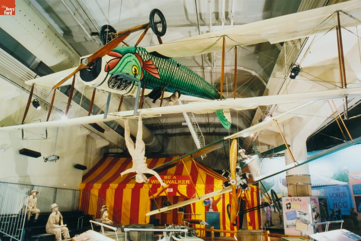 1917 Curtiss JN-4D "Canuck" Biplane Plane with white wings and body painted like a green-scaled fish, hanging upside from ceiling in front of yellow-and-red striped tent