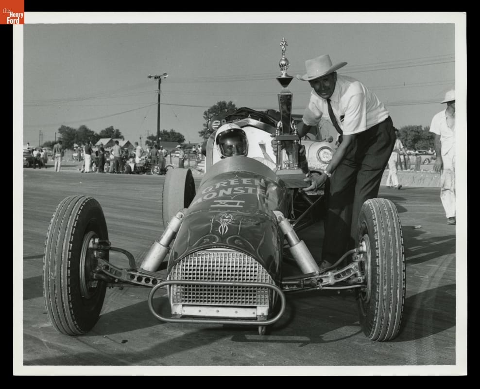 Art Arfons and Wally Parks with the Trophy for Top Speed, NHRA Nationals, Detroit Dragway, 1959 Man in cowboy hat bends down to hand large trophy to man in helmet in race car