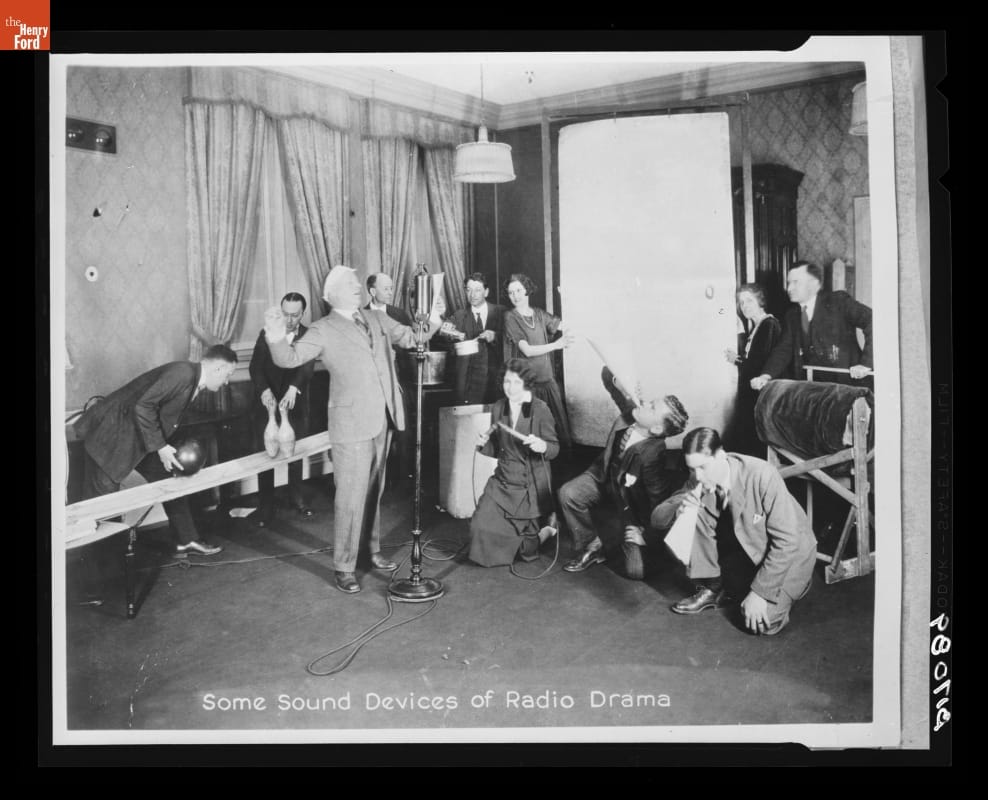 Man at a microphone is surrounded by many people with various noise-making items
