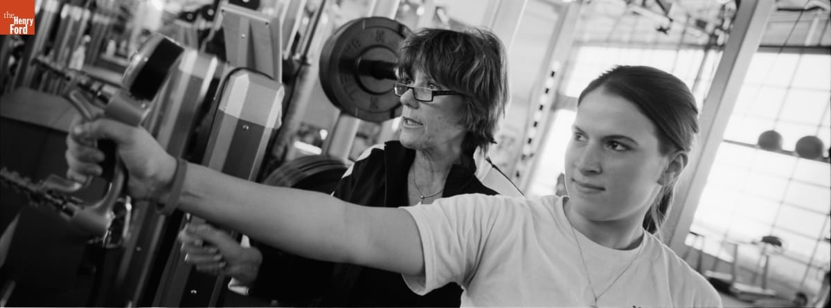 Two women, one holding a piece of equipment in her hand, stand in a gym looking leftward
