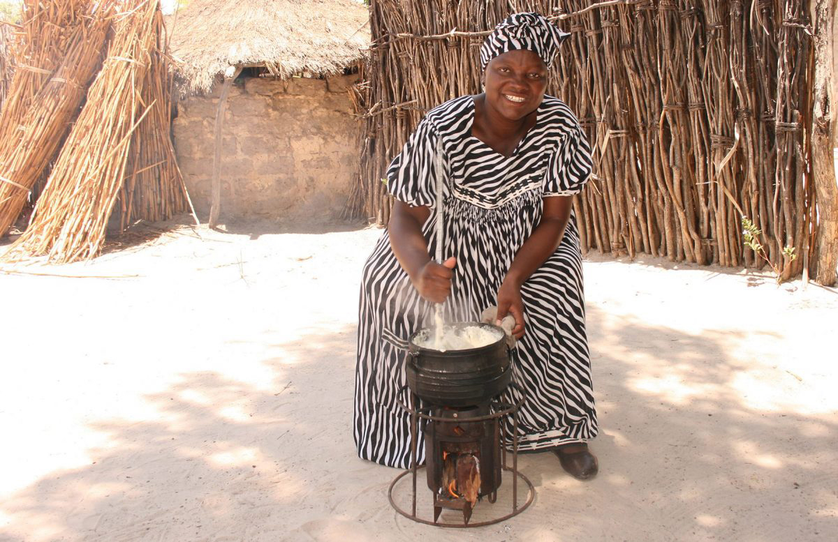 Dark-skinned woman in black-and-white zebra print dress and headwrap stirs a pot over a portable stove in front of a wall of reeds or sticks