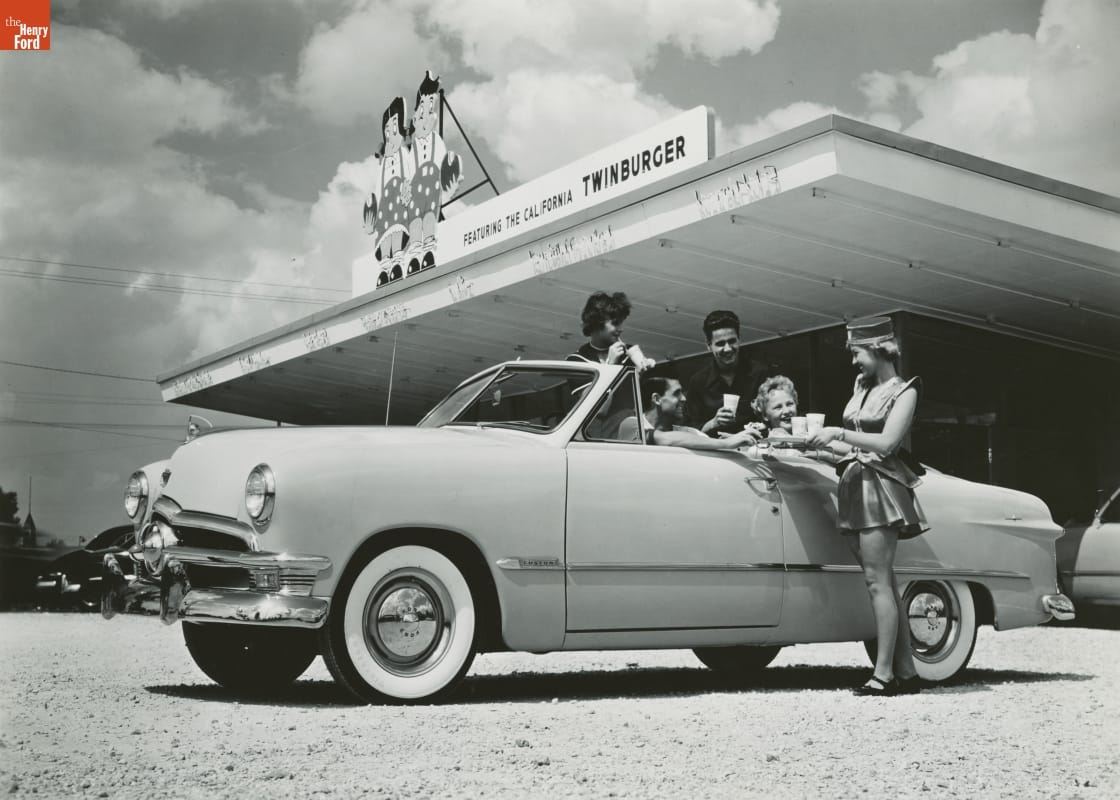 Black-and-white photo of a group of young adults in a convertible in front of a restaurant; a female carhop holds a tray by the car