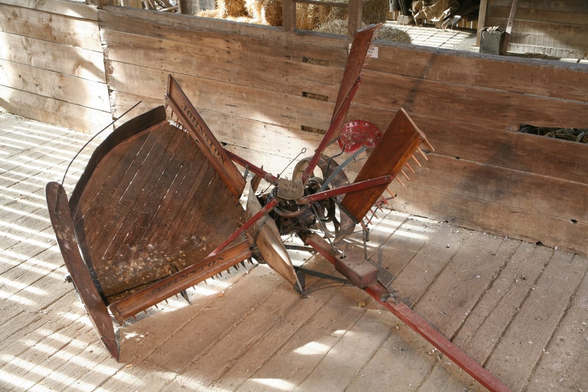 Piece of large farm equipment, with wooden platform, long wooden beam, and toothed wooden rakes, sitting in wooden barn