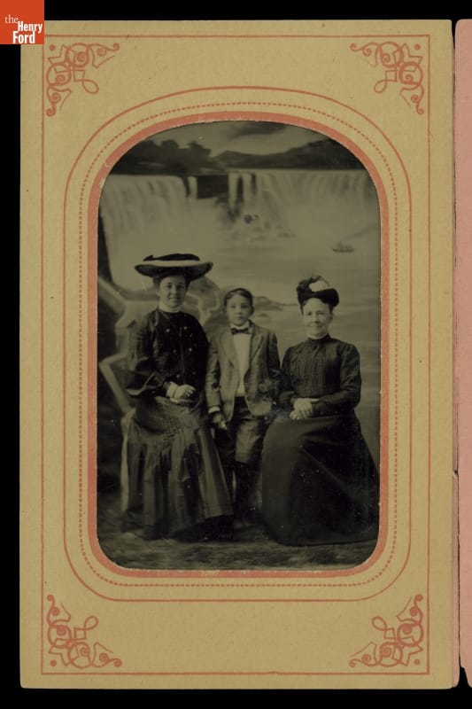 Two seated women in dresses and hats with small boy standing between them, all in front of a painted backdrop of a waterfall