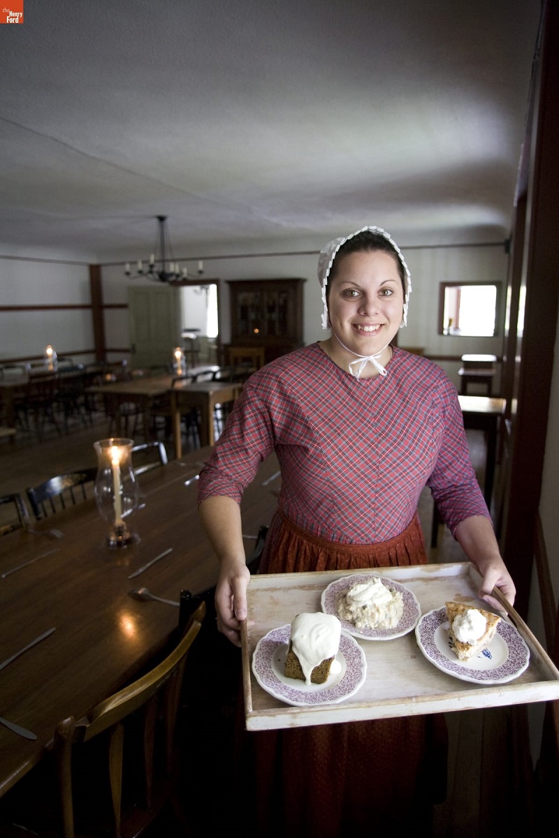 Eagle Tavern in Greenfield Village, October 2007 / Photograph by Michelle Andonian Smiling woman in pink dress and white bonnet holds a tray of food in a large room with low ceilings and long wooden tables