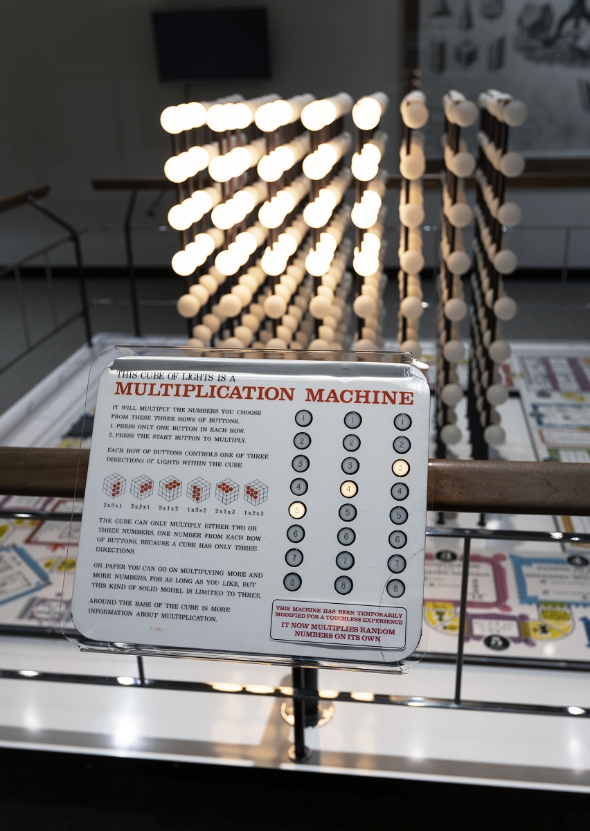 Lightbulbs arranged in a cube shape, some lit and some not, behind a wood and metal railing with a large sign in the foreground containing text