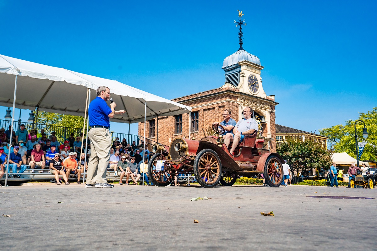 Man in a blue shirt and khakis holding a microphone gestures to an old-fashioned open car with two people in it, as spectators look on from the side