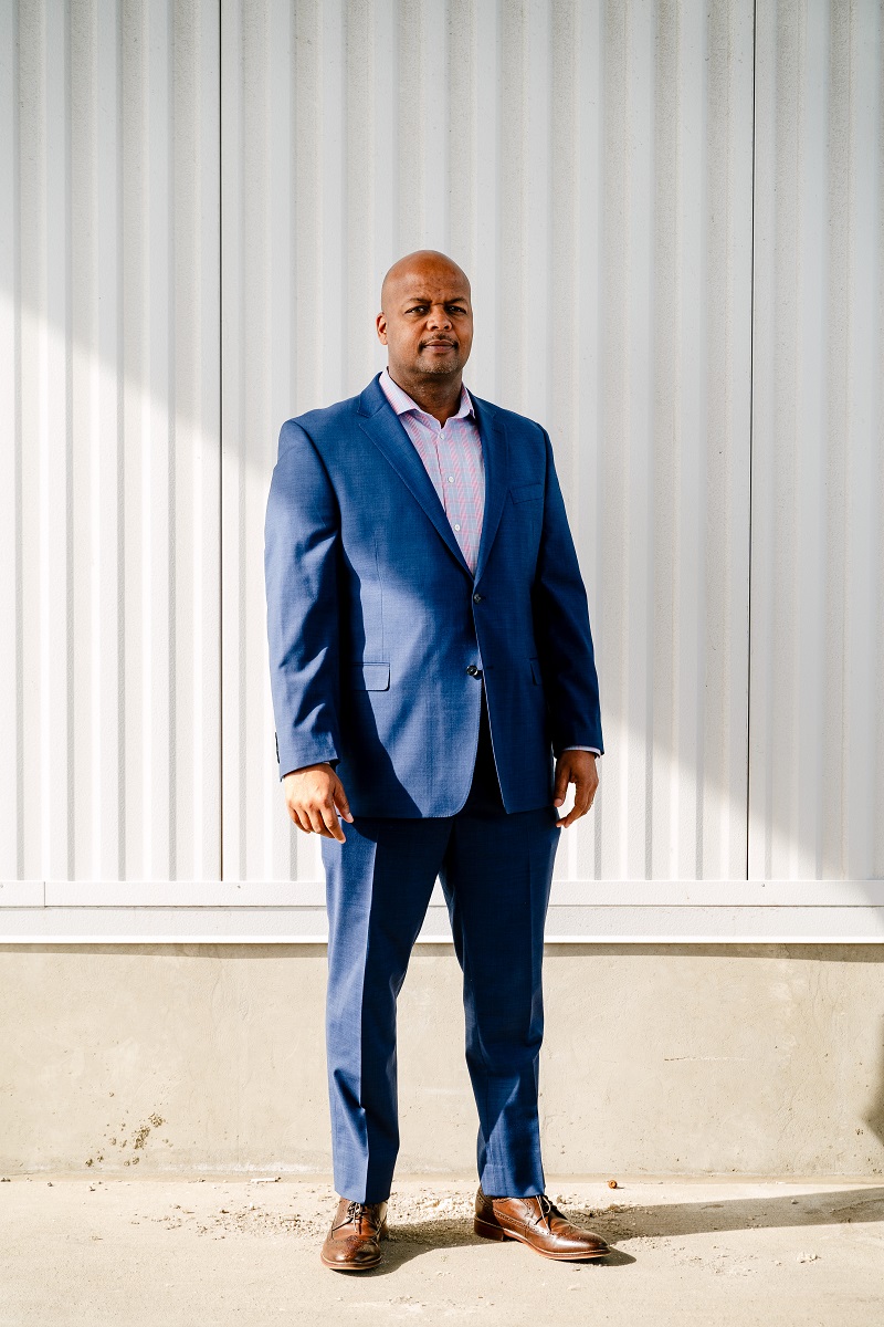 Corey Williams, Dearborn Truck Plant Manager at Ford Motor Company Medium-skin tone man in a blue suit stands in front of a corrugated white metal wall