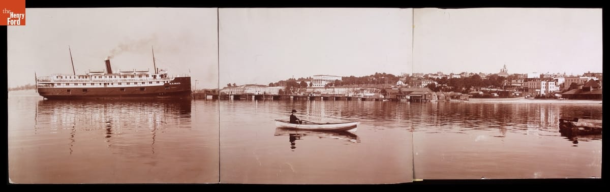 Man in a rowboat paddles near a larger steamer in a harbor in front of docks and a town
