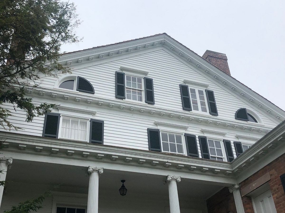 Peaked gable of a white wooden house with rectangular and quarter-circle-shaped windows; above a porch (?) with columns