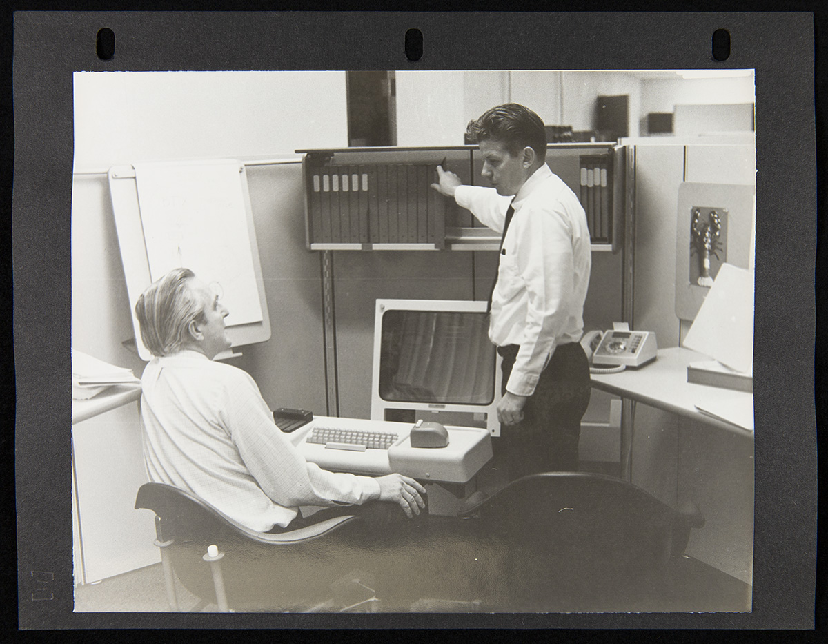Two men, one seated and one standing, among office furniture holding a computer, phone, and books or binders