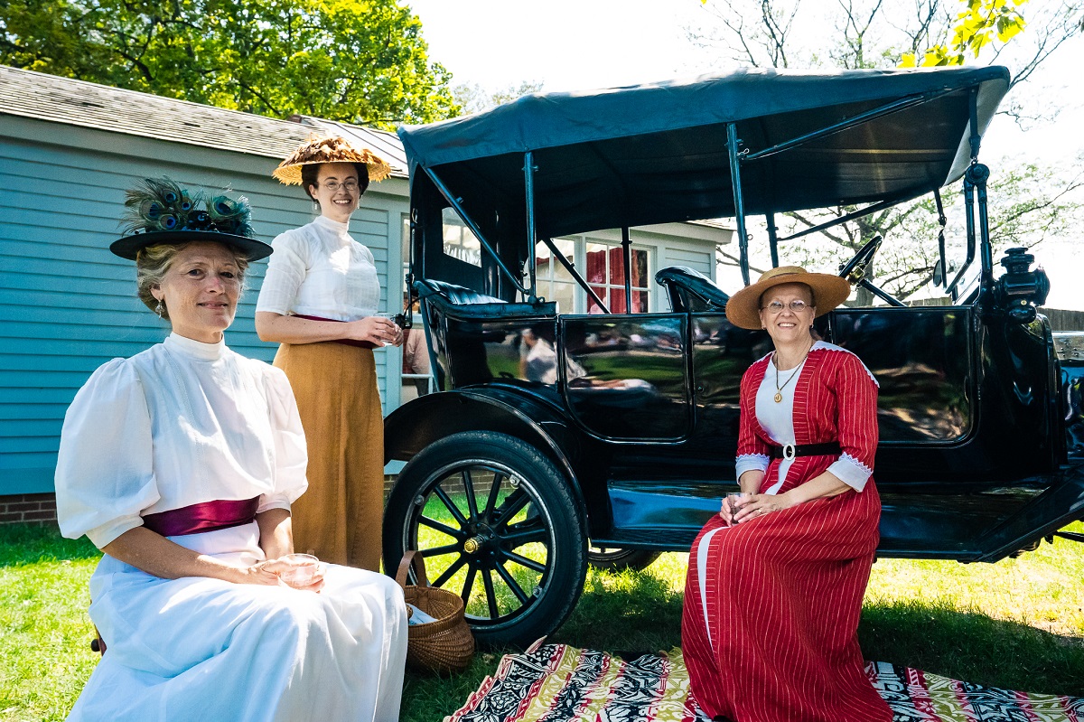 Three women in historic clothing and hats sit and stand by/on a black car