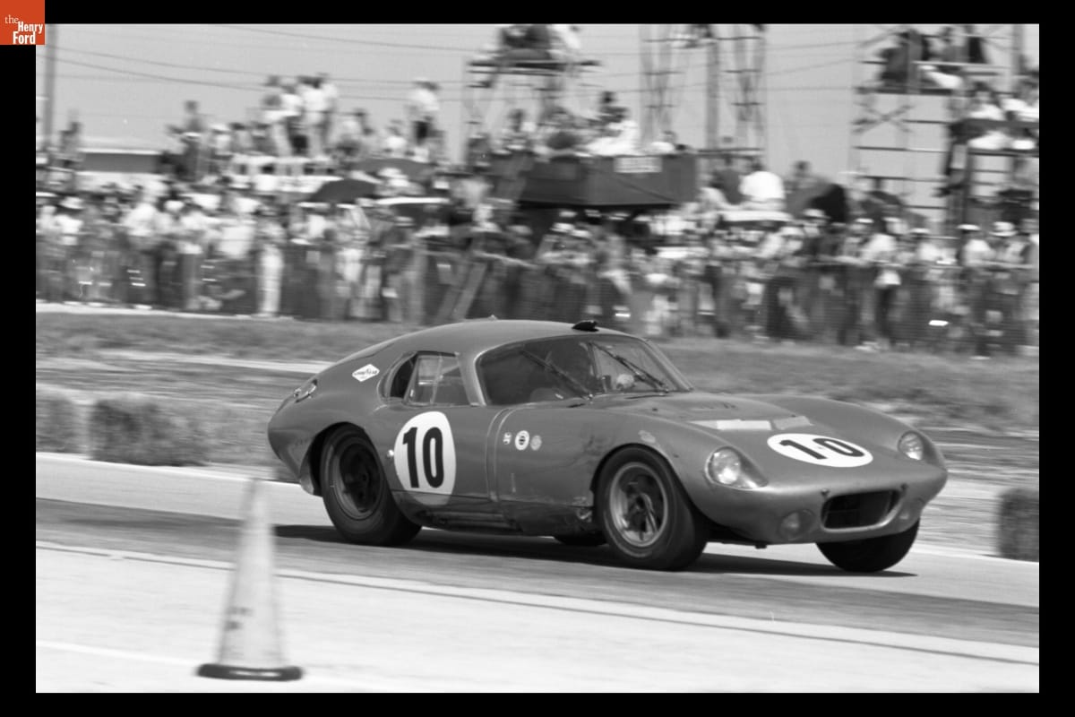Black-and-white photo of low racecar with long hood on a track in front of fans