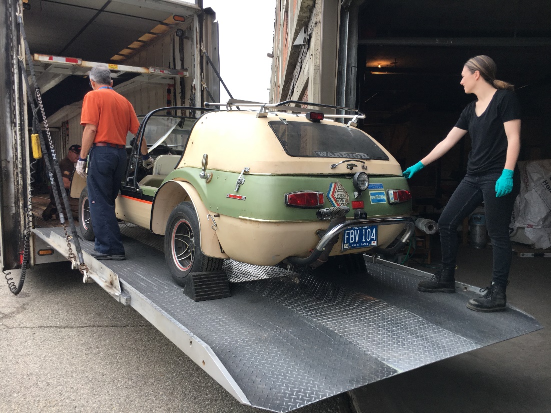 Two people stand with an open beige-and-green car on the loading ramp of a truck; one kneels in front