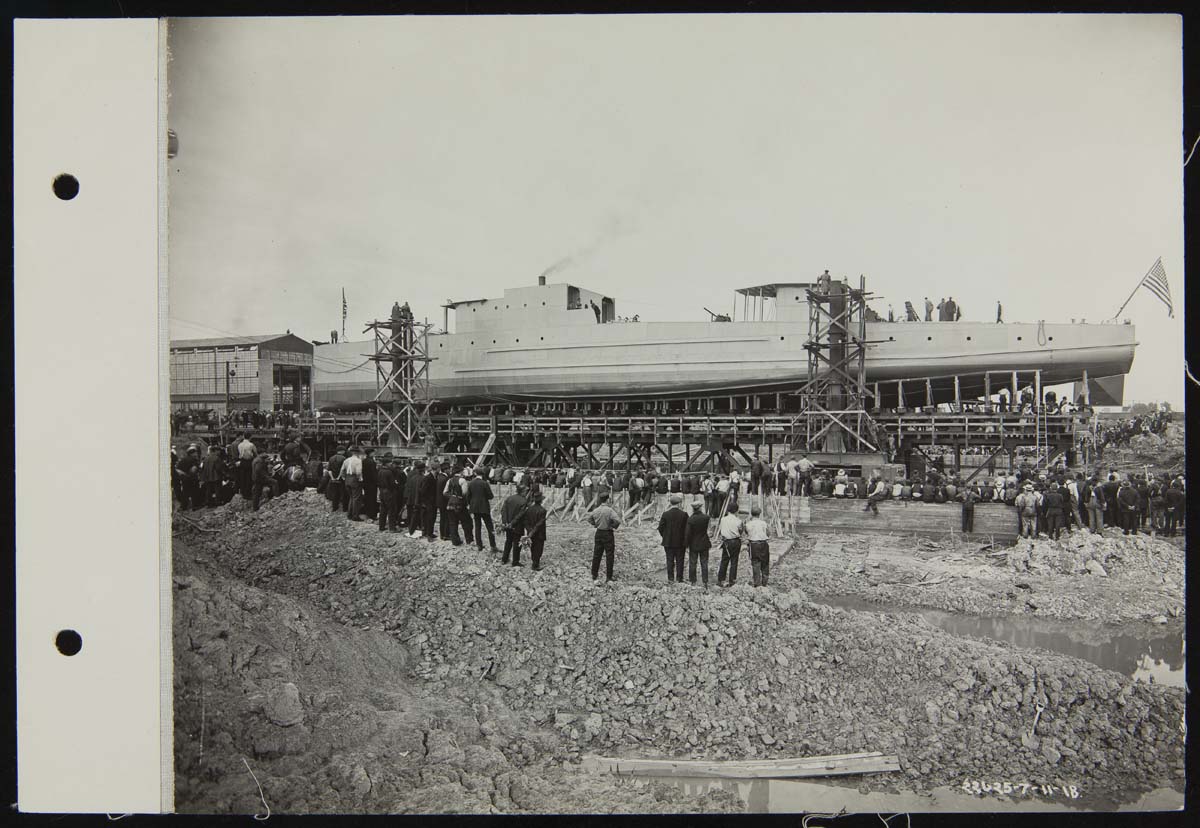 Long, narrow boat on elevated trestle with a large group of people on land looking on